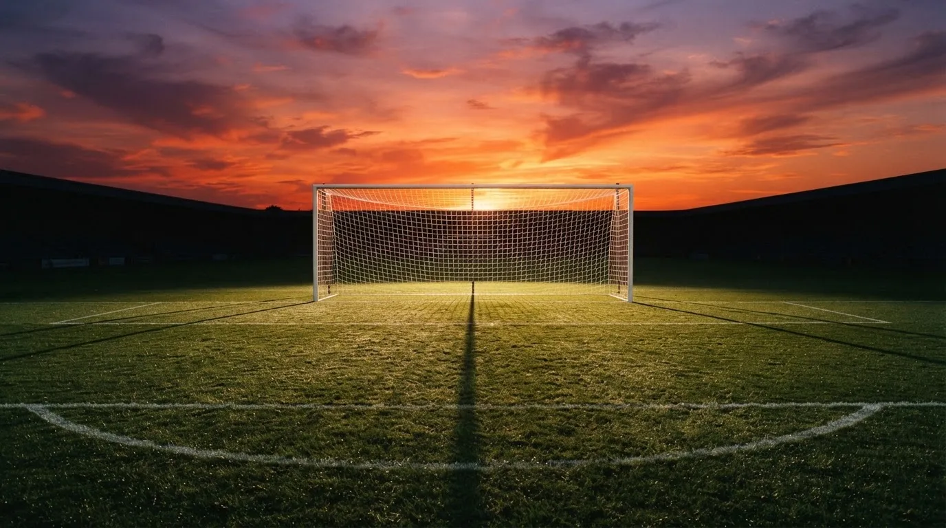 Ein Fußballtor mit Netz bei Sonnenuntergang auf einem leeren Platz, die Schatten der Torpfosten liegen lang auf dem Gras.
