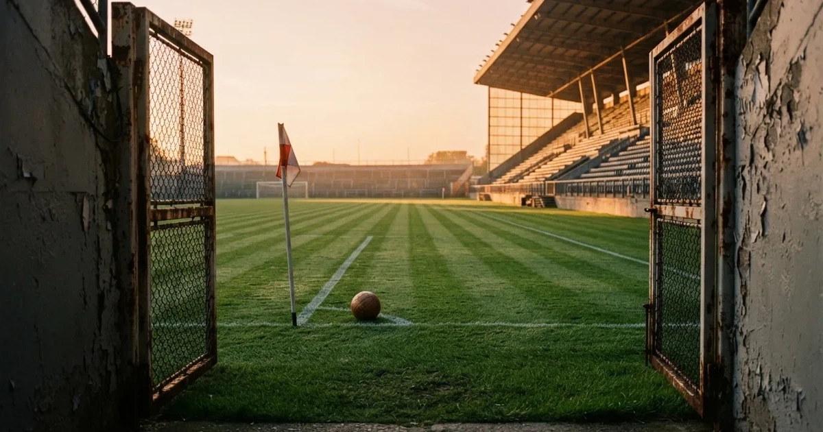 Blick durch ein Stadiontor auf einen grünen Rasen im Abendlicht, ein Ball ruht an der Eckfahne, die Tribünen sind leer.