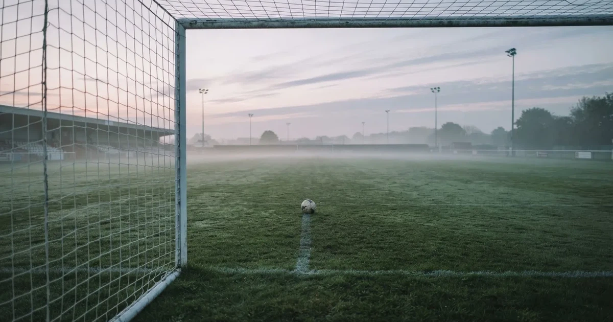 Ein Fußballtor mit gespanntem Netz steht auf einem leeren Rasenplatz, davor ein einzelner Ball, Morgendunst liegt über dem Feld.