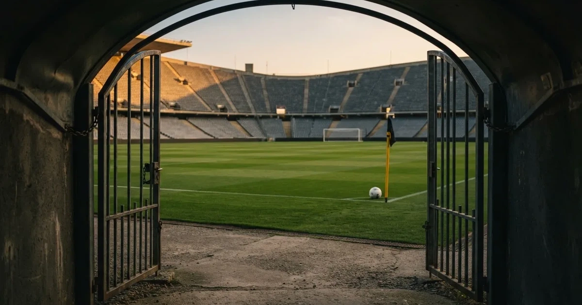 Blick durch ein Stadiontor auf einen grünen Rasen im Abendlicht, ein Ball liegt an der Eckfahne, die Ränge sind leer.