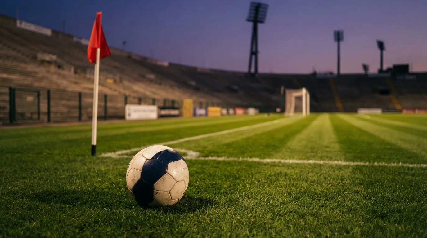 Ein Fußball liegt neben einem Eckfahnen-Pfosten auf einem Rasenplatz, im Hintergrund ein leeres Stadion in der Dämmerung.