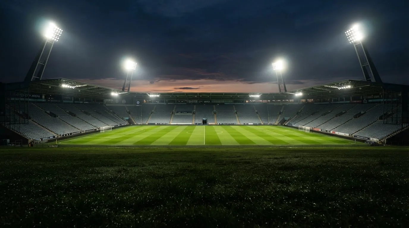 Eine leere Stadiontribüne unter Flutlicht aus der Ferne betrachtet, die Rasenfläche leuchtet grün in der Dämmerung.