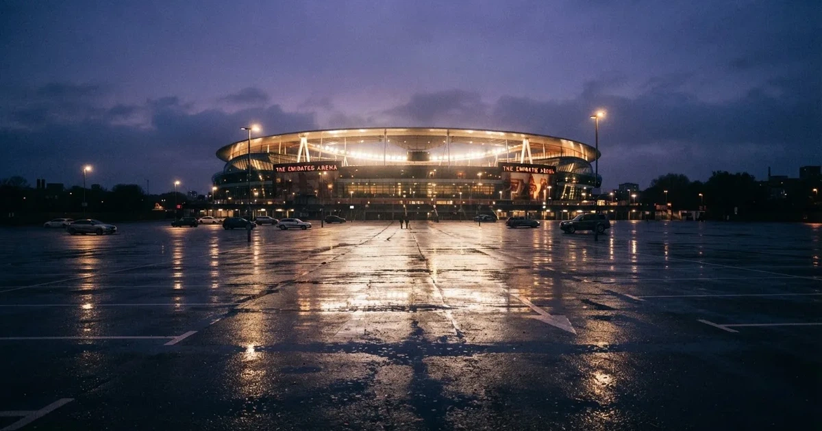 Ein Stadion unter Flutlicht aus großer Entfernung gesehen, davor ein regennasser Parkplatz in der Dämmerung.