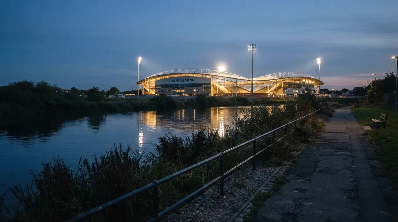 Ein Stadion unter Flutlicht aus der Ferne gesehen, im Vordergrund ein ruhiger Fluss und ein Fußweg.