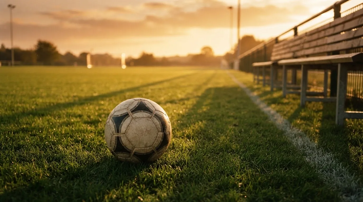 Ein Fußball auf einem Rasenplatz im Gegenlicht der tiefstehenden Sonne, am Rand eine leere Ersatzbank.