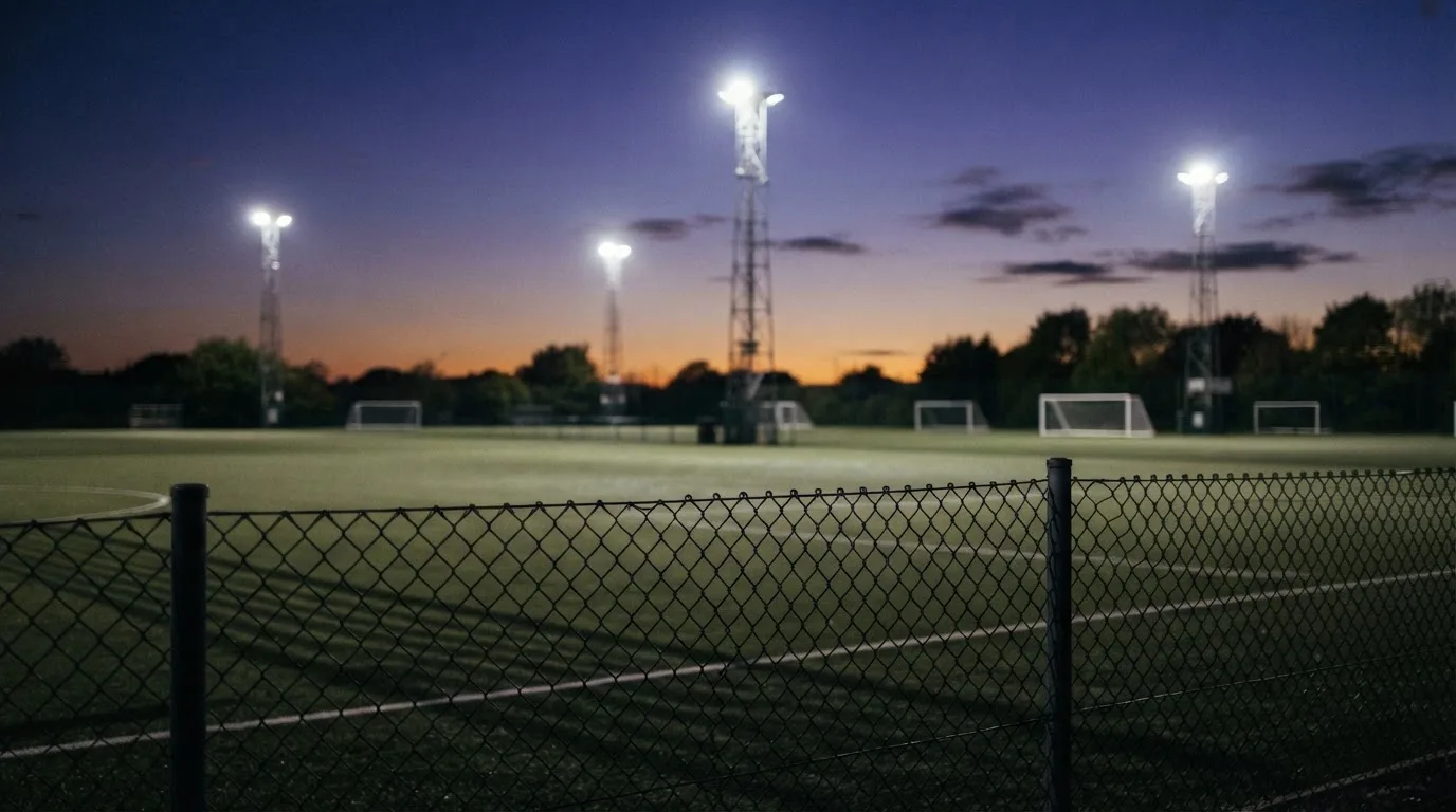 Eine Flutlichtanlage über einem leeren Sportplatz am Abend, im Vordergrund ein Maschendrahtzaun.