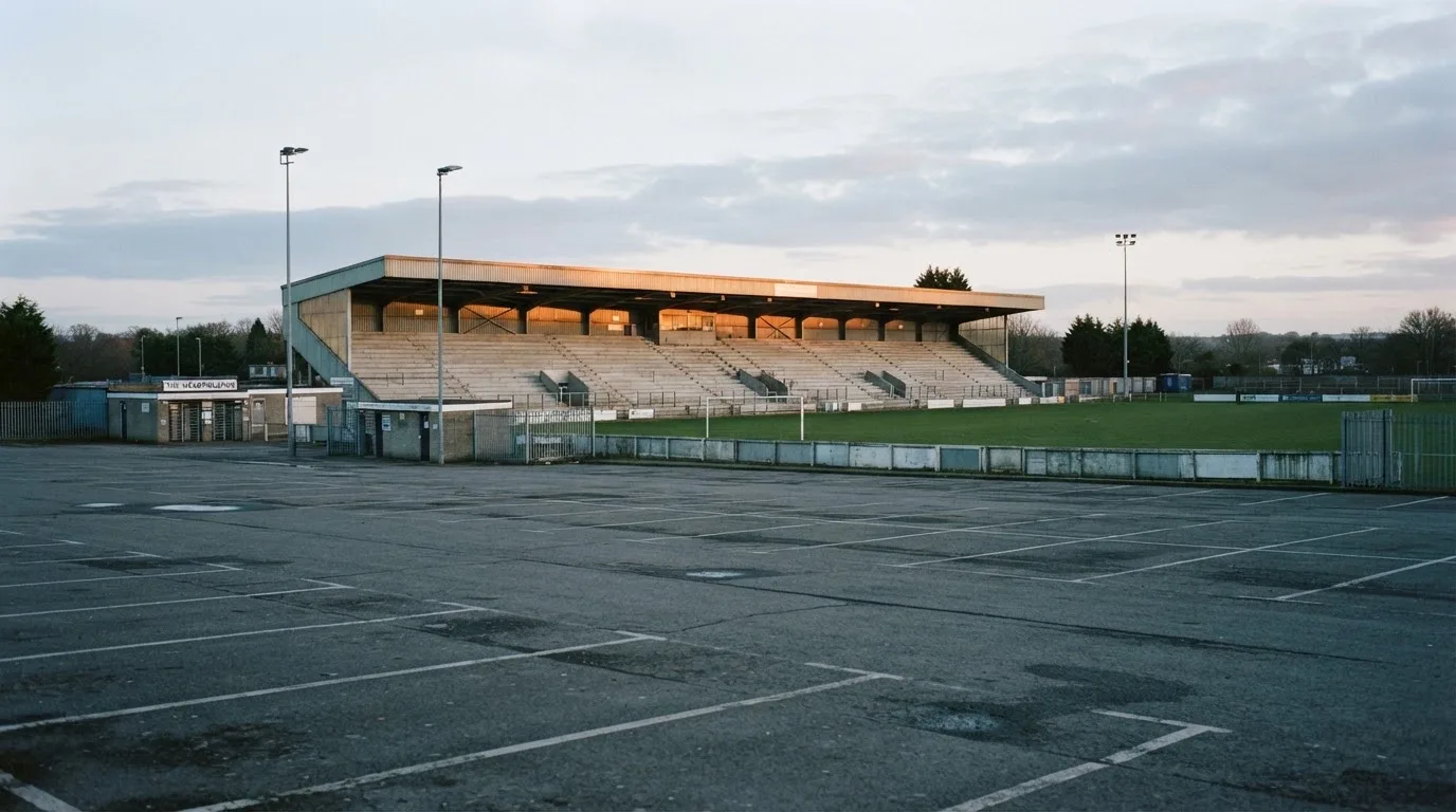 Eine leere Tribüne eines kleinen Stadions am frühen Morgen, aus der Ferne über einen menschenleeren Parkplatz fotografiert.