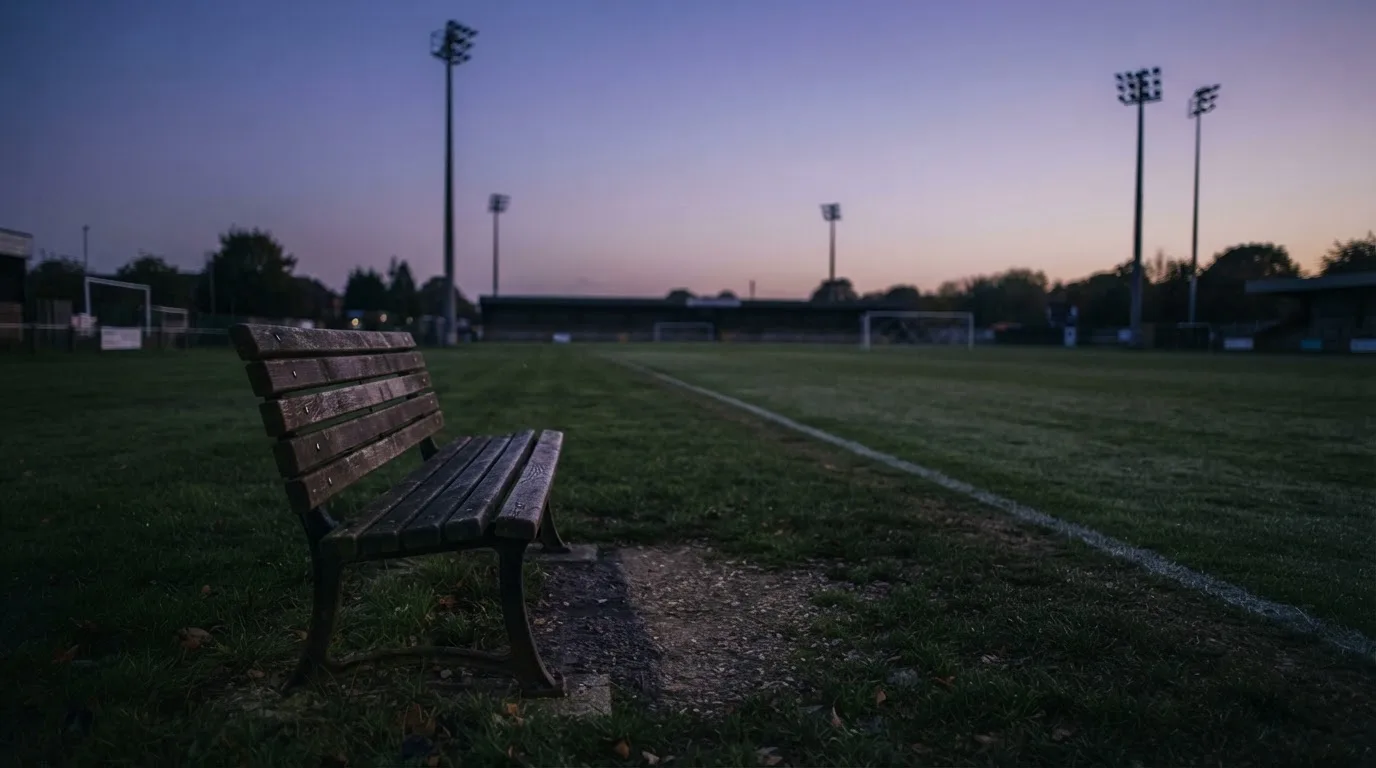 Eine leere Parkbank am Rand eines Fußballfelds in der Abenddämmerung, im Hintergrund ragen dunkle Flutlichtmasten in den Himmel.