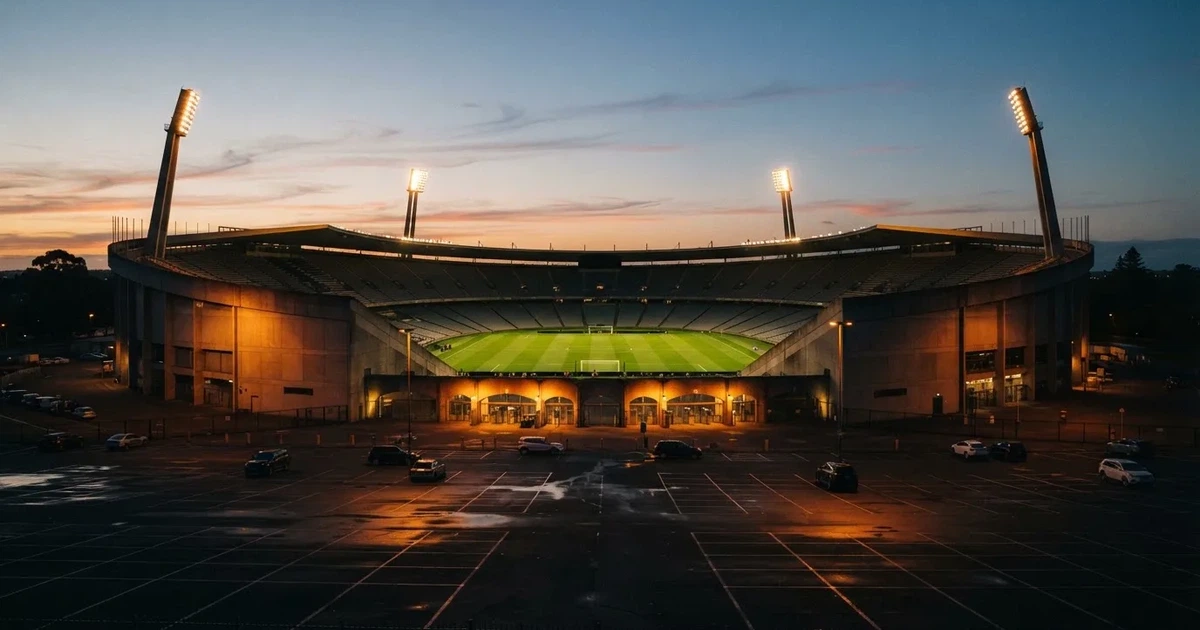 Ein Fußballstadion unter Flutlicht aus großer Entfernung, davor ein dunkler Parkplatz und ein heller Abendhimmel.