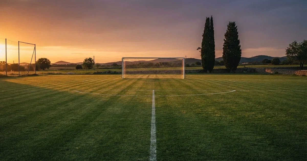 Ein Fußballtor mit Netz auf einem leeren Platz im Abendlicht, der Rasen frisch gemäht, im Hintergrund Zypressen.