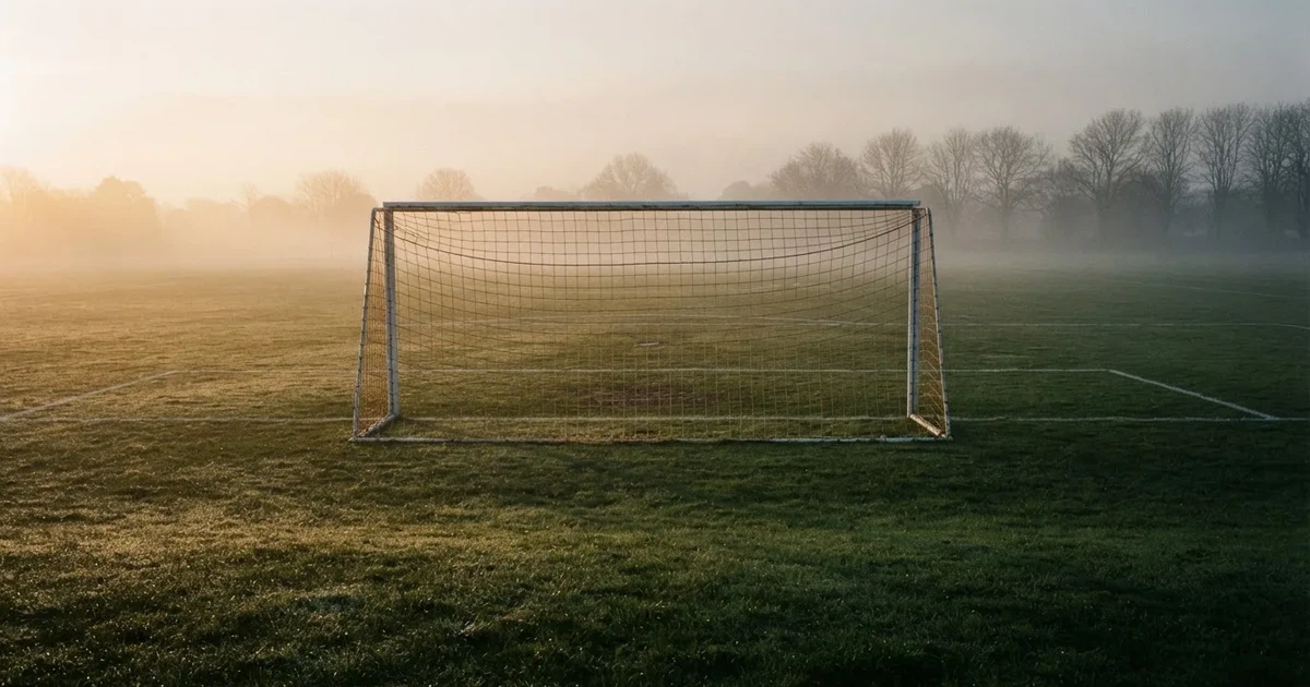 Ein Fußballtor mit Netz steht auf einem leeren Platz im Nebel der Morgendämmerung, der Rasen ist taufeucht.