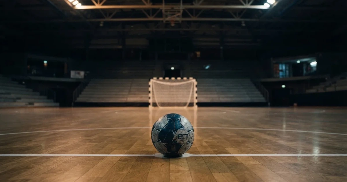 Ein Handball liegt auf dem Hallenboden vor einem Tor mit Netz, die leere Halle wird von Flutlicht beleuchtet.