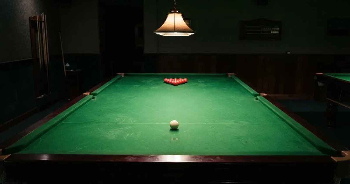 Ein Snookertisch mit grünem Tuch und aufgebauten roten Kugeln in einem abgedunkelten Raum unter einer hängenden Lampe.