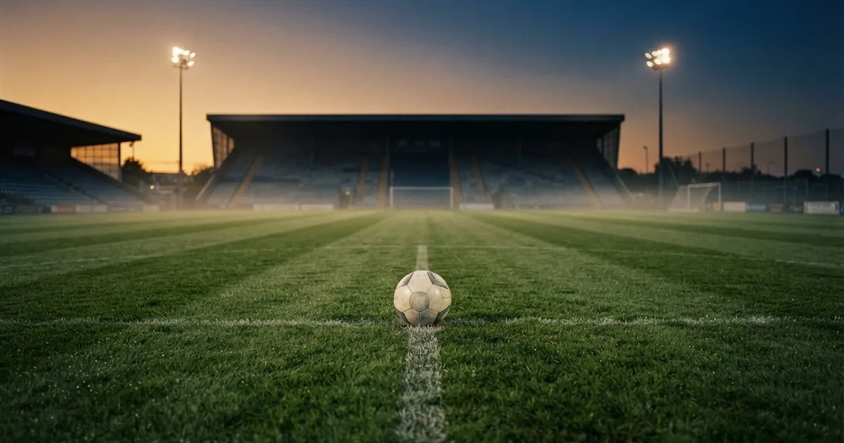 Ein Fußball liegt auf dem Anstoßpunkt eines leeren Rasenplatzes, im Hintergrund eine Tribüne und Flutlichtmasten in der Abenddämmerung.