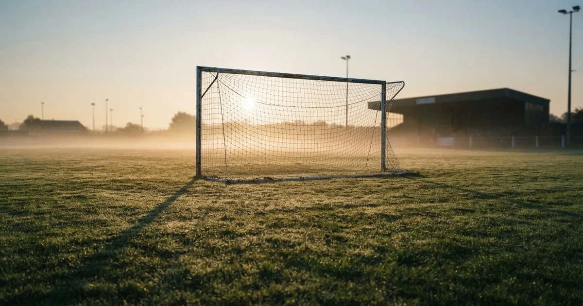Ein Fußballtor mit Netz steht im Morgennebel auf einem leeren Rasenplatz, im Hintergrund die Silhouette einer kleinen Tribüne.