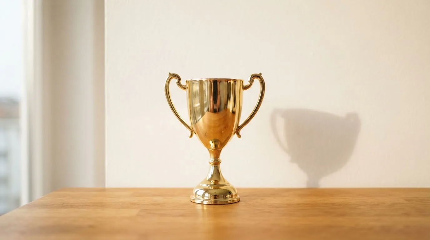 Ein Pokal ohne Gravur steht allein auf einem Holztisch vor einer hellen Wand, davon fällt ein weicher Schatten.