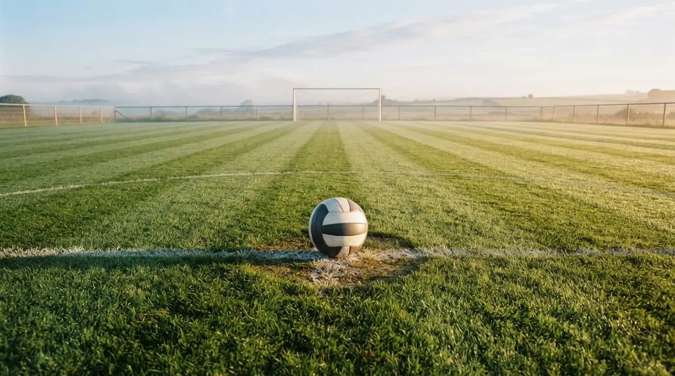 Ein Fußball liegt allein auf dem Anstoßpunkt eines leeren Rasenplatzes im Morgenlicht, am Rand ein niedriger Zaun.