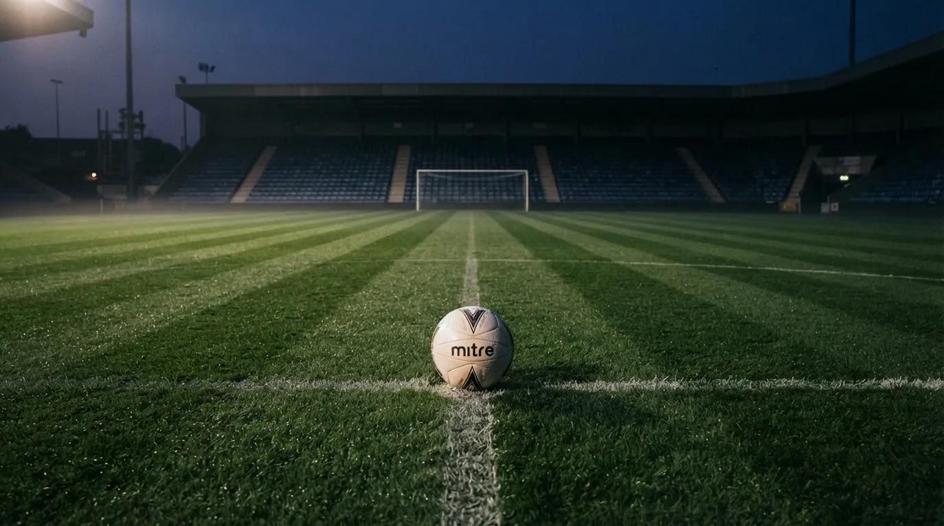Ein Fußball liegt auf dem Anstoßpunkt eines leeren Rasenplatzes, dahinter ragen dunkle Ränge in den Abendhimmel.