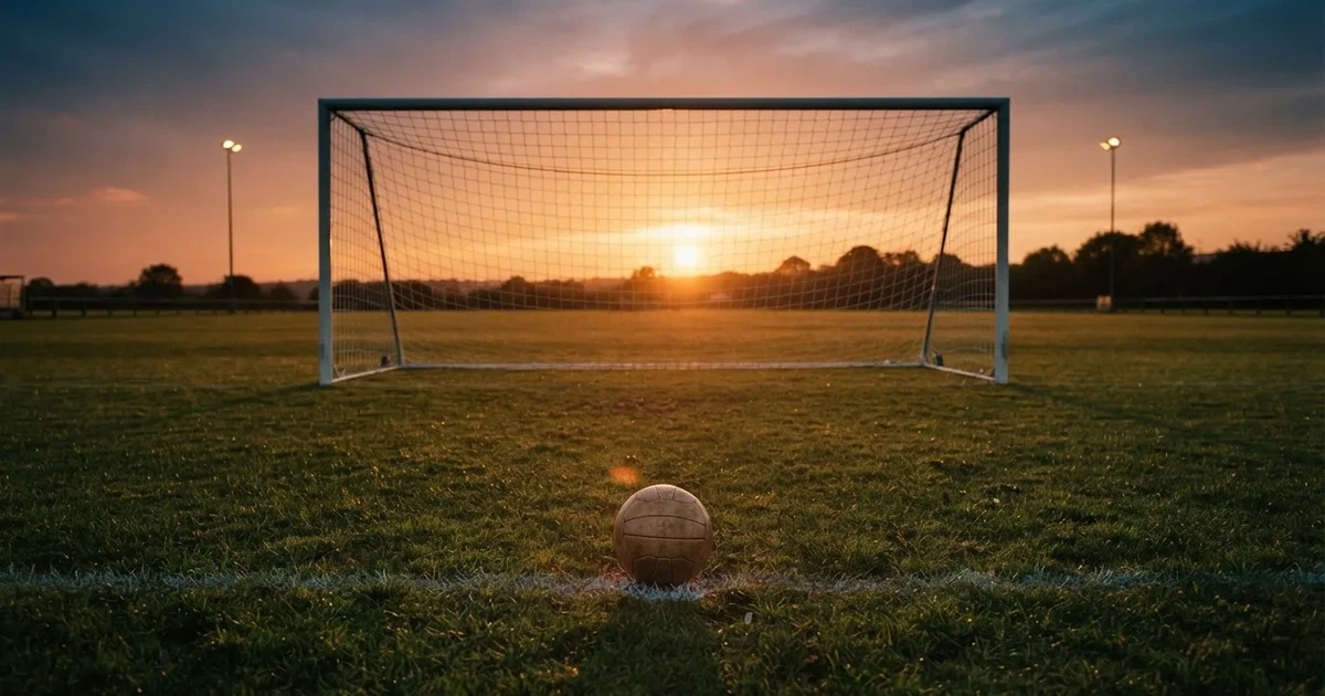 Ein Fußballtor mit Netz im Gegenlicht auf einem leeren Platz am Abend, davor ein Ball auf dem Rasen.