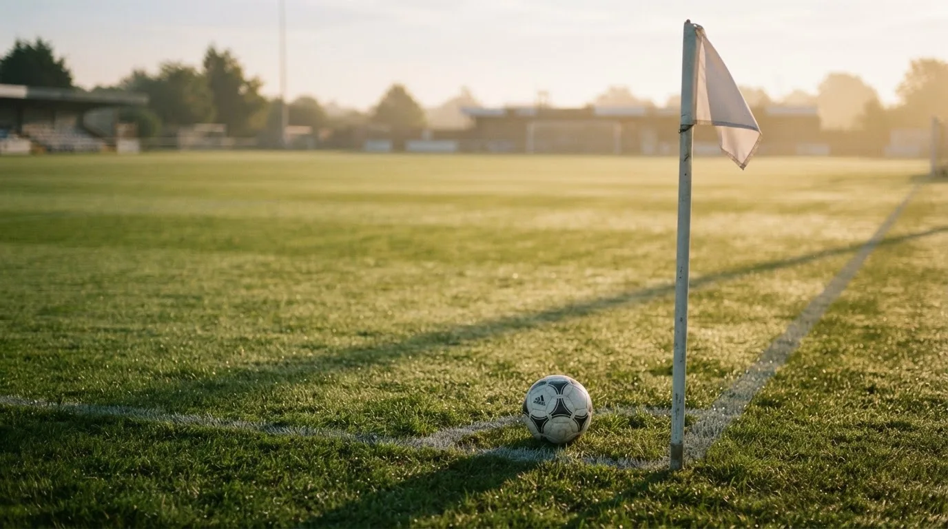 Ein Fußball liegt auf Rasen neben einer Eckfahne ohne Aufdruck auf einem leeren Platz im Morgenlicht.
