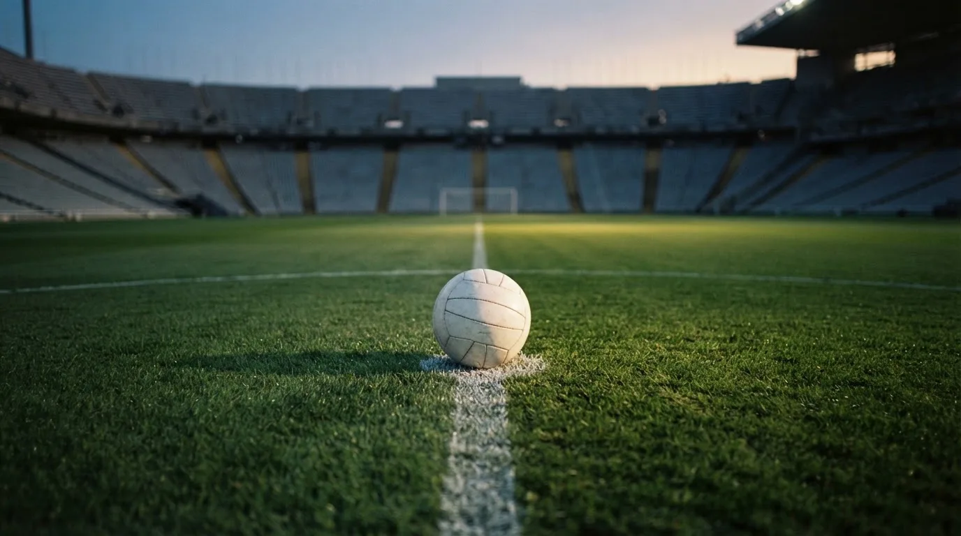 Ein Fußball liegt auf dem Anstoßpunkt eines Rasenplatzes, dahinter eine leere Tribüne im Abendlicht.