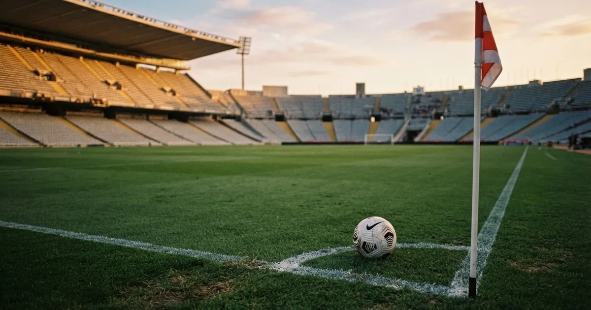 Ein Fußball liegt am Viertelkreis an der Eckfahne eines Rasenplatzes, im Hintergrund leere Tribünen in warmem Abendlicht.