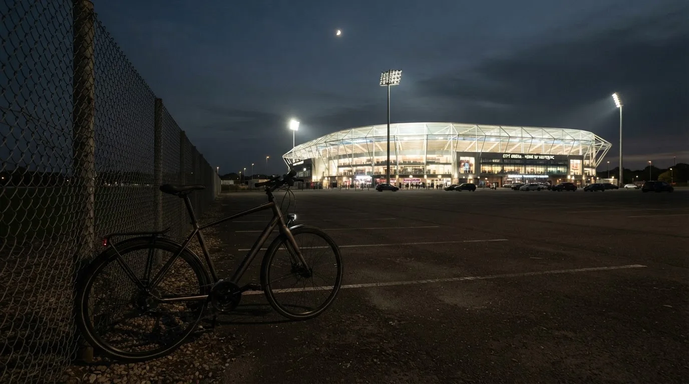 Ein Stadion unter Flutlicht aus der Ferne gesehen, davor ein dunkler Parkplatz und ein einzelnes Fahrrad an einem Zaun.