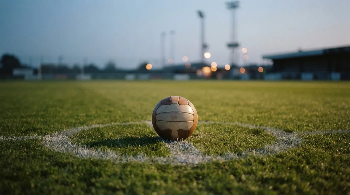Ein Fußball liegt auf dem Anstoßpunkt eines Rasenplatzes in der Abenddämmerung, Flutlichtmasten stehen unscharf im Hintergrund.