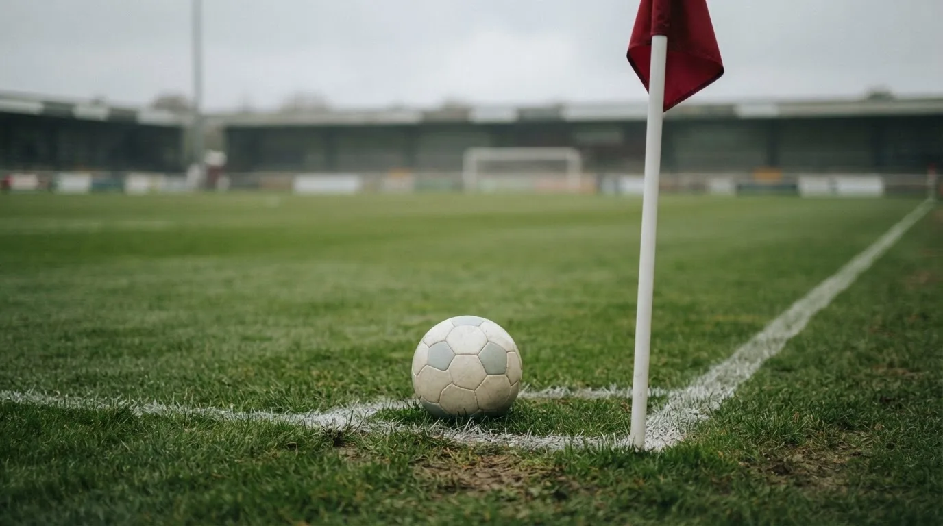 Ein Fußball liegt neben einer Eckfahne auf kurz gemähtem Rasen unter bewölktem Himmel.